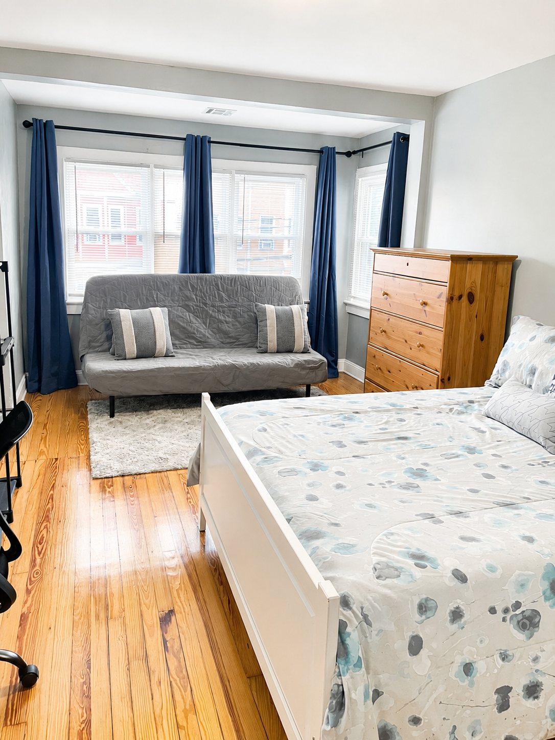 Bright bedroom with a bed, gray futon, wooden dresser, and blue curtains