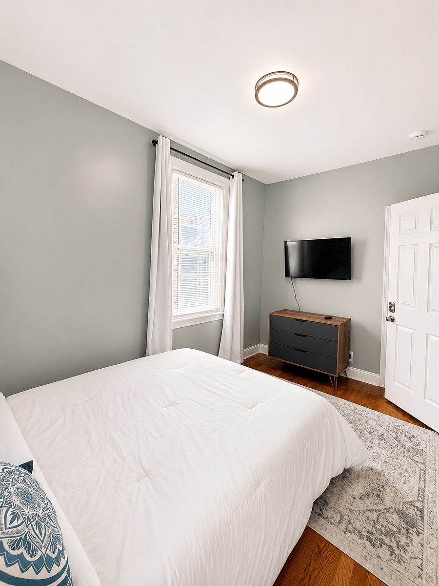 Furnished bedroom with white bedding, dresser, and wall-mounted television