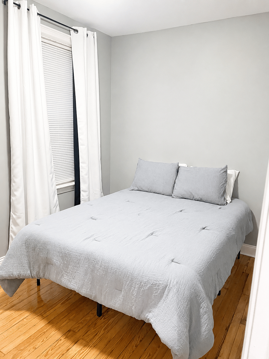 Furnished bedroom with gray bedding beside a curtained window